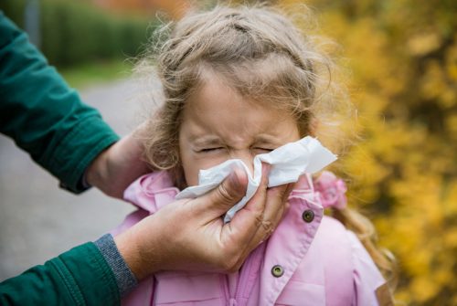 Father wiping daughter's nose with handkerchief. Sick little girl with cold and flu standing outdoors. Preschooler sneezing, coughing, having runny red nose. Autumn street background